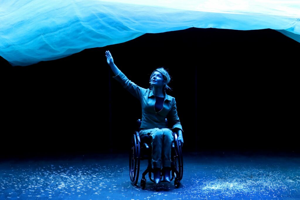 Photo de Dorine dans son fauteuil qui veut toucher du doigt une grande vague bleue au-dessus d'elle